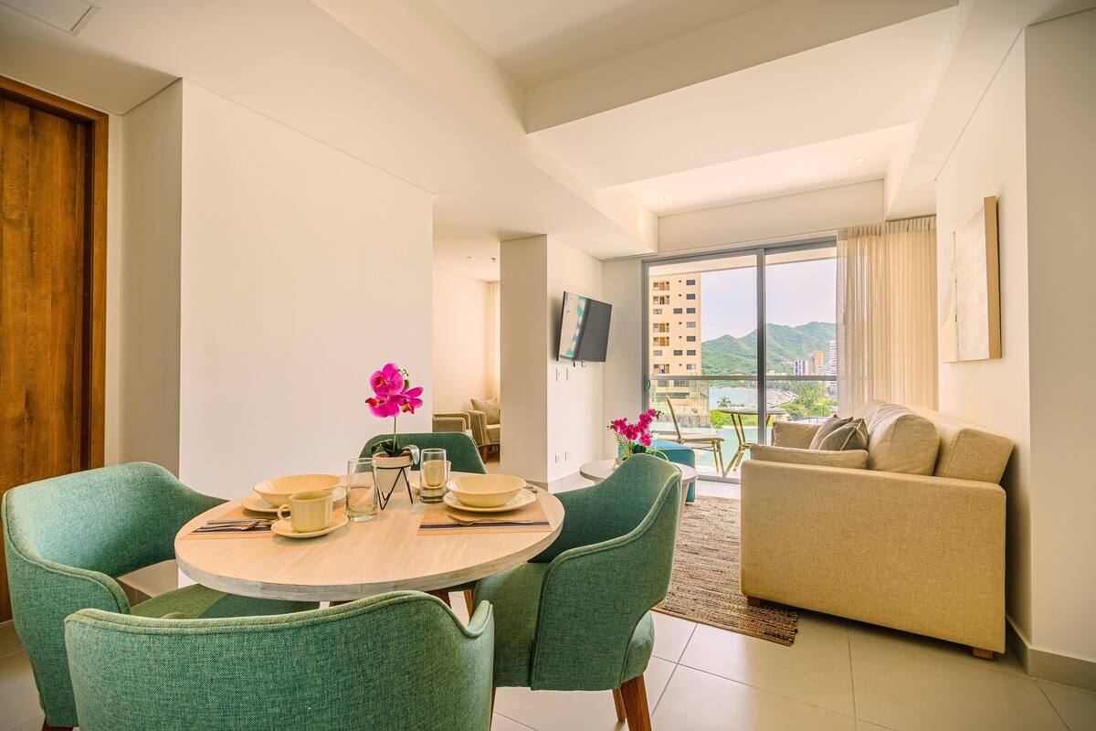A modern living room with a round dining table, green chairs, a beige sofa, and a view of mountains through large windows.