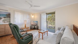 Uma sala de estar ampla com uma poltrona de couro verde, um sofá cinza, uma mesa de centro de madeira e uma televisão sobre um suporte. Há uma janela grande com cortinas, um ventilador de teto e um quadro na parede.
