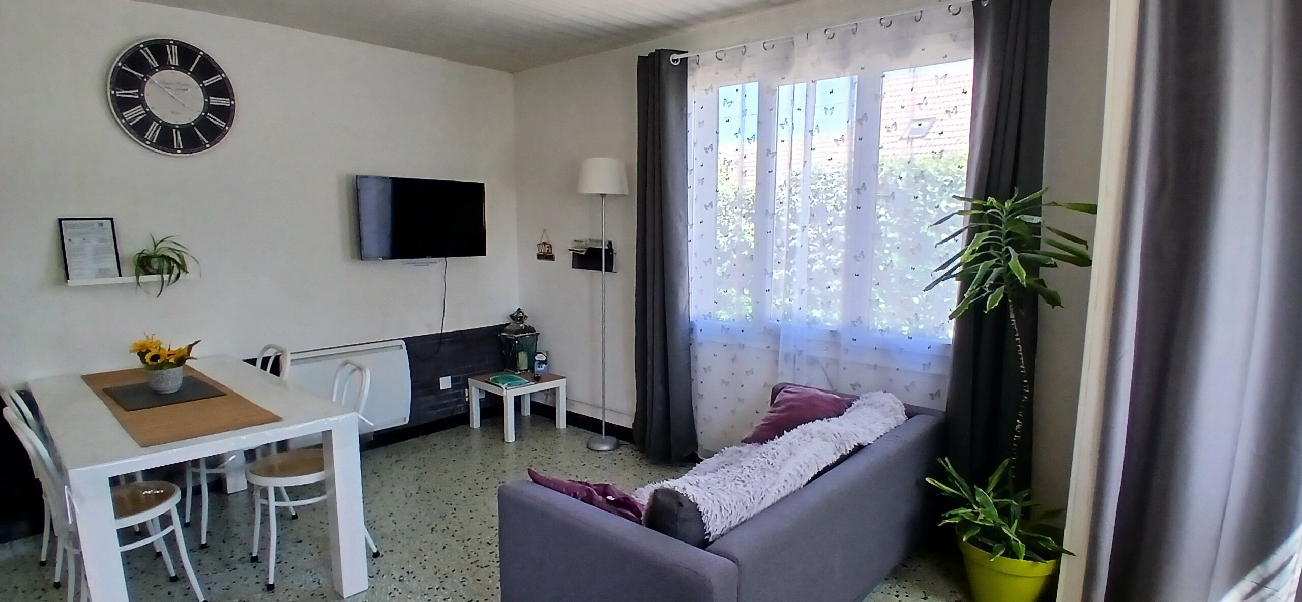 Ein Wohnzimmer mit einer Couch, einem Esstisch mit Stühlen, einem an der Wand befestigten Fernseher und einem Fenster mit Vorhängen.