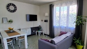 Una sala de estar con un sofá, una mesa de comedor con sillas, un televisor montado en la pared y una ventana con cortinas.