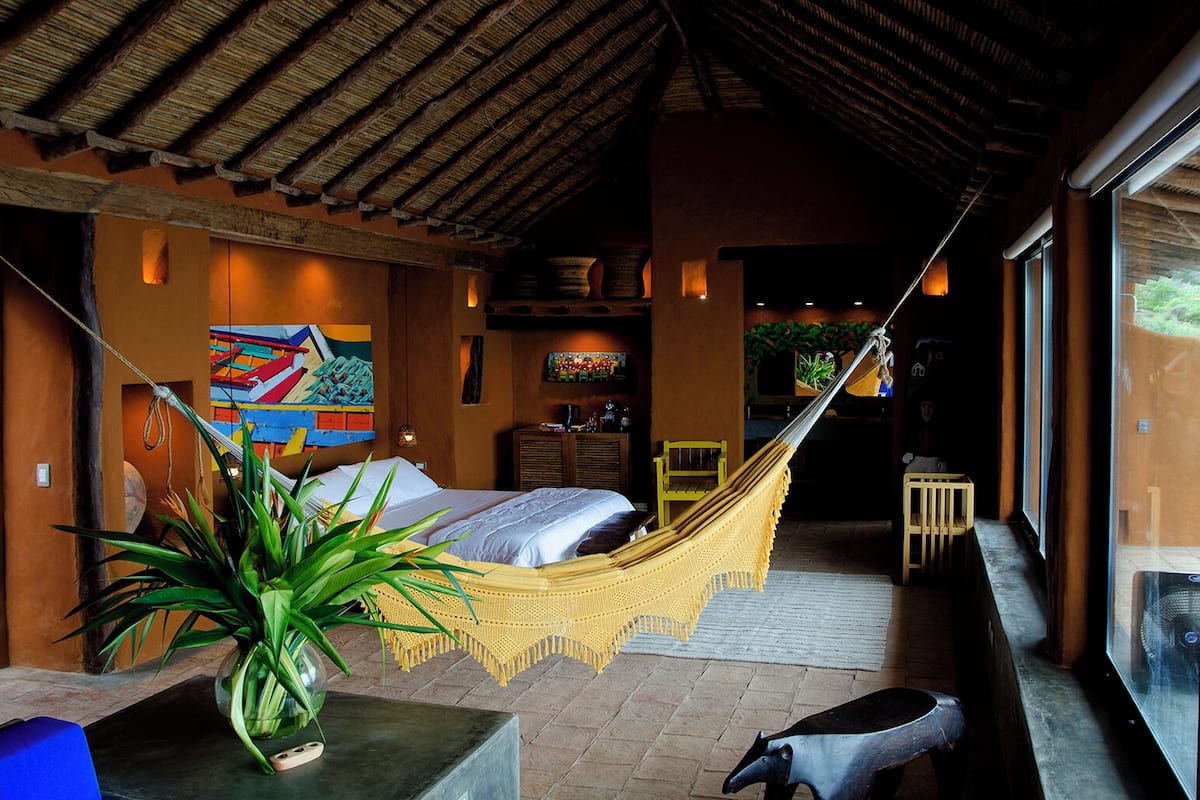 Una habitación con una cama, una hamaca, una silla, una mesa y un ventanal grande.