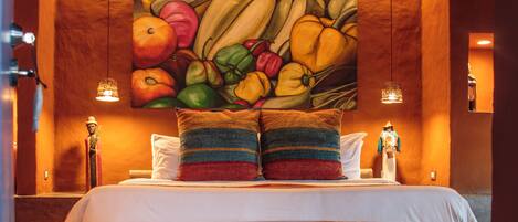 Una cama con cabecera de madera, un cuadro de bodegón con frutas encima y dos lámparas fijadas a la pared.