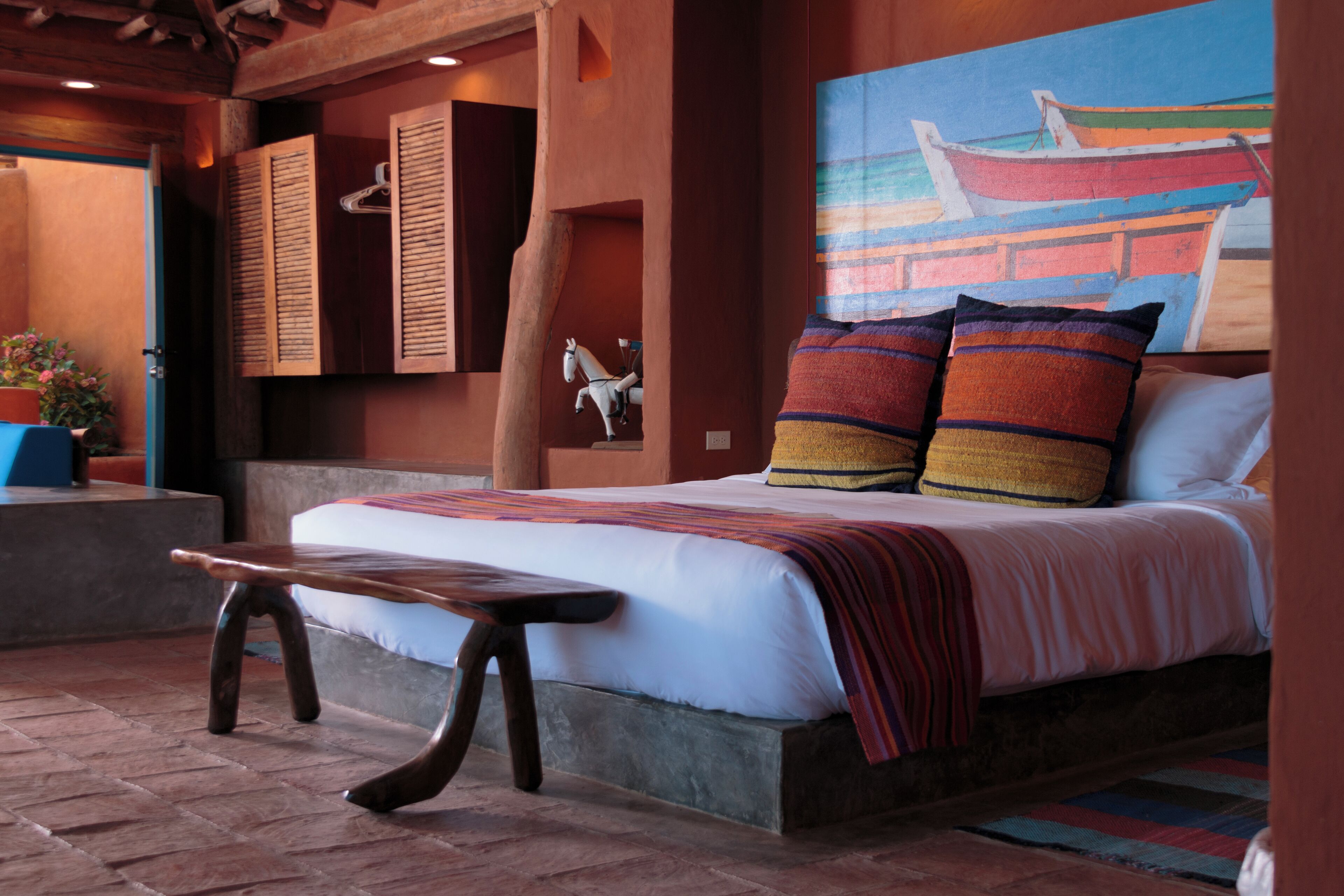 A bedroom with a bed, pillows, a bench, and a painting of a boat on the wall.