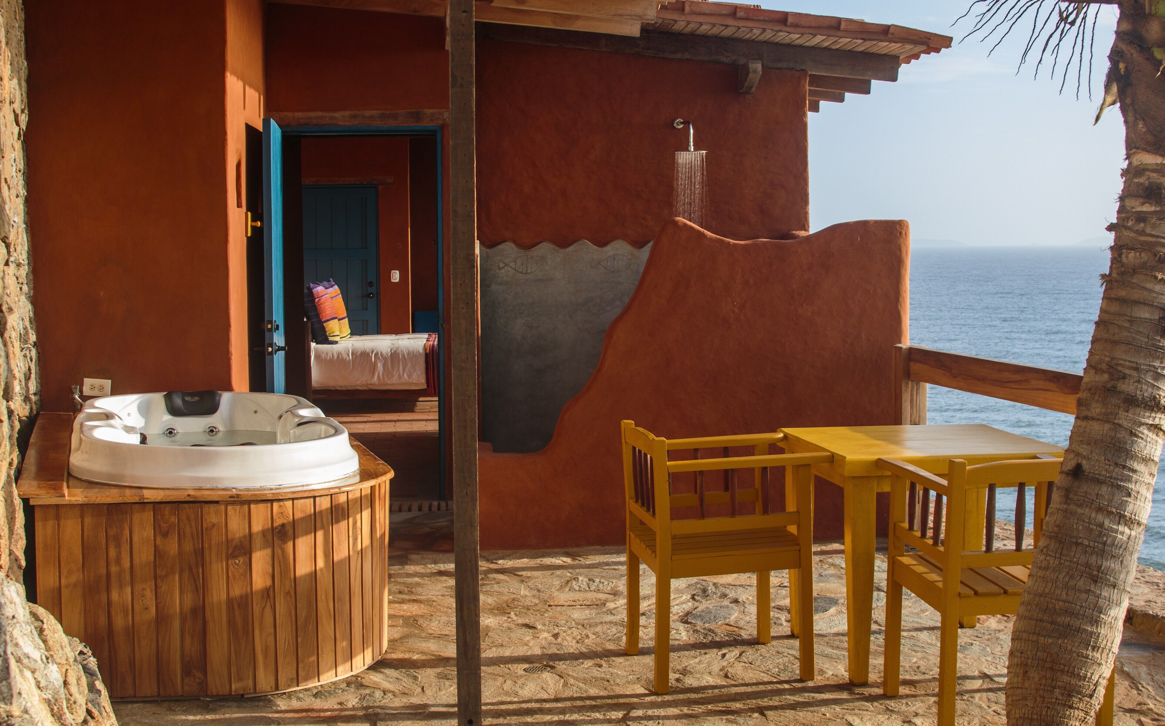 A wooden hot tub, a wooden table, and chairs on a stone patio with a view of the ocean.