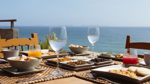 Een gedekte tafel met verschillende ontbijtproducten, glaasjes water en sap, en uitzicht op de oceaan.