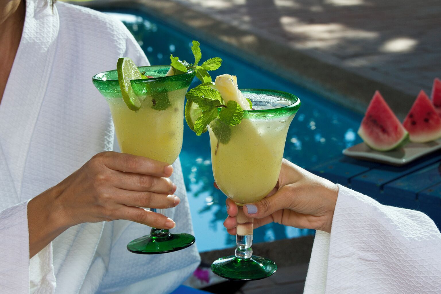 Dos personas sosteniendo vasos de una bebida amarilla con adorno de lima y menta junto a una piscina.
