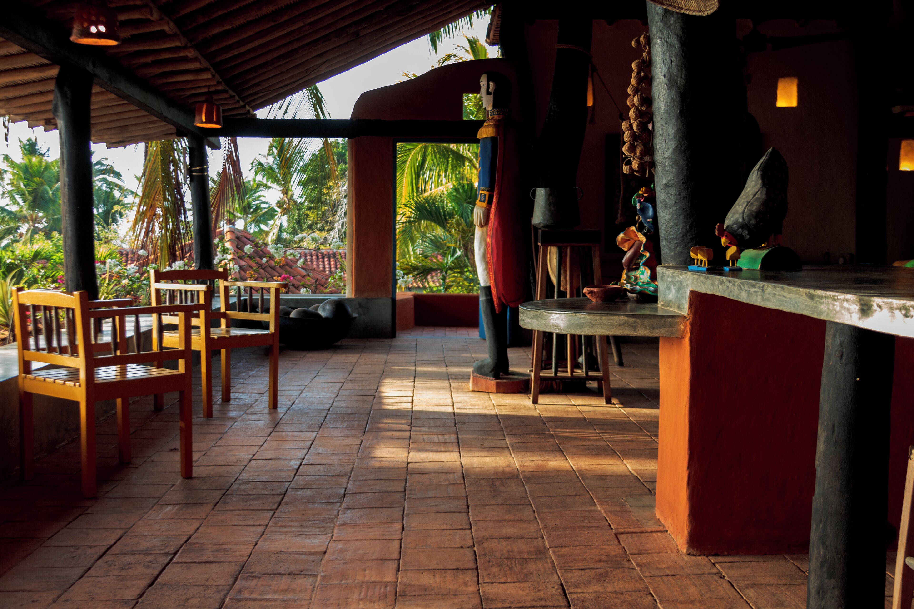 A covered outdoor seating area with wooden chairs and a table, surrounded by palm trees and a tiled floor.