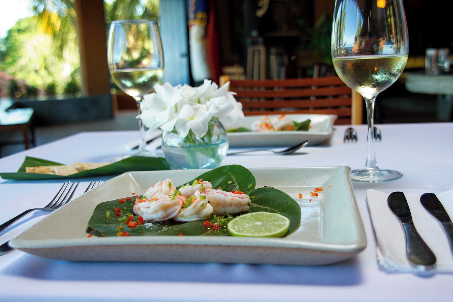 A dining table with plates of food, a glass of white wine, and a bowl of flowers.
