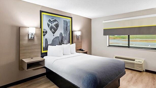 Una habitación de hotel moderna con una cama grande, mesitas de noche, una lámpara de pared, una ventana con vistas a la carretera y un aire acondicionado instalado en la pared.