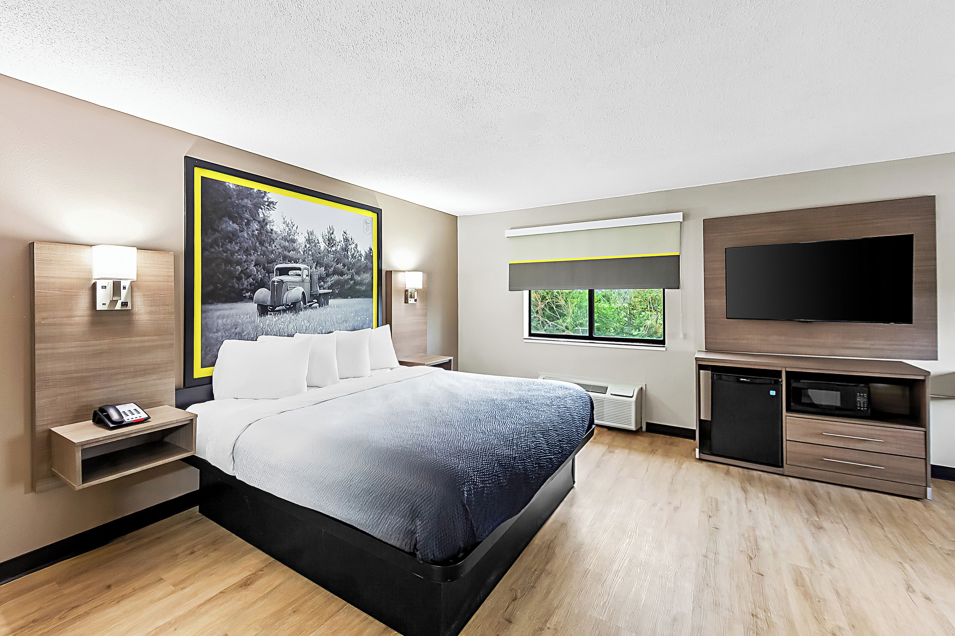 A modern hotel room with a bed, bedside table with a phone, a TV, a microwave, and a large window with a view of greenery.