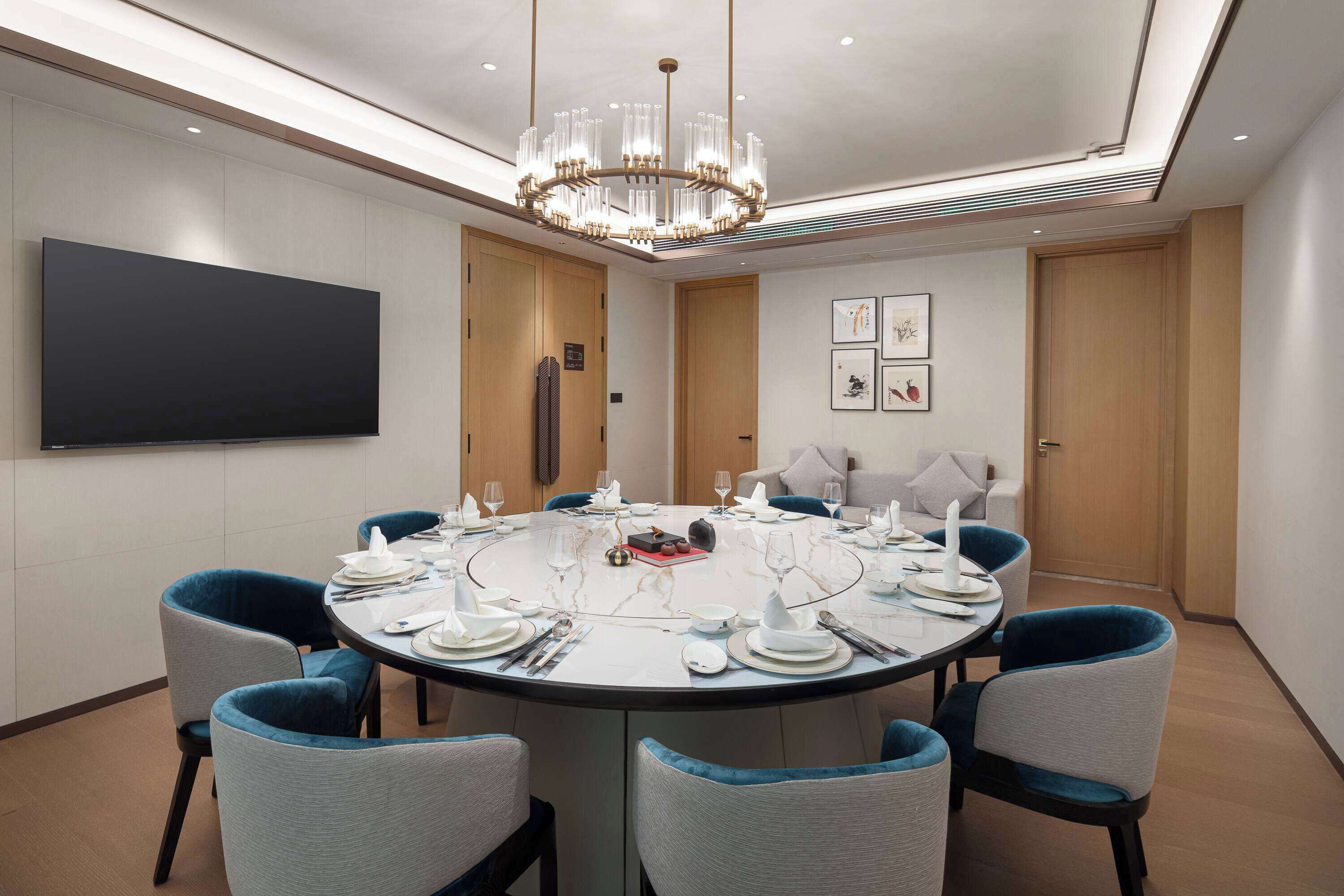 Une salle de conférence avec une table ronde préparée pour une réunion, entourée de chaises, d’un grand téléviseur à écran plat et d’un lustre moderne.