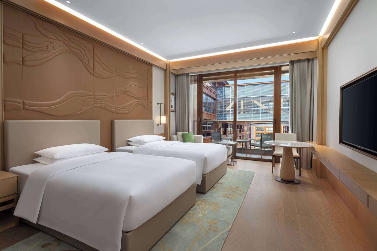 A hotel room with two beds, a desk, a chair, a TV, and a balcony with a view of the city.
