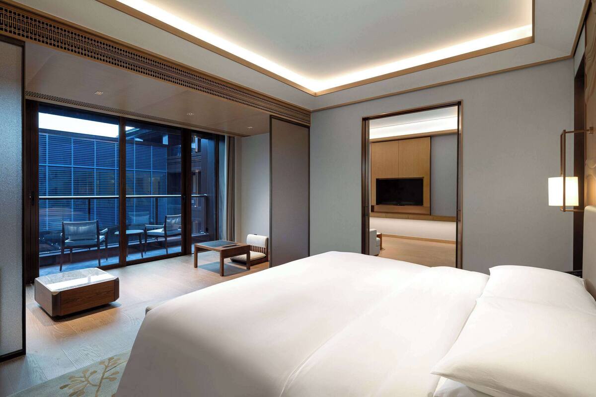 A modern hotel room with a large bed, a TV, a balcony with seating, and a sliding door leading to an outdoor area.