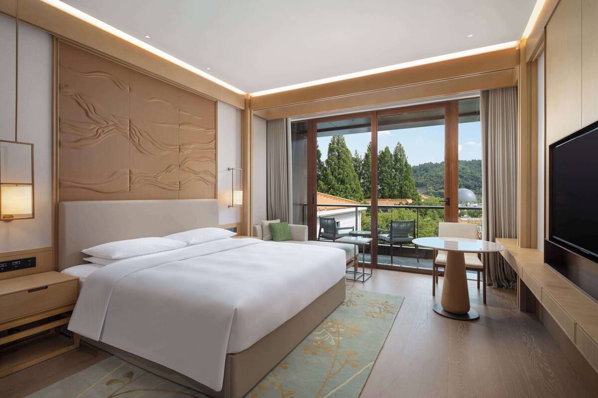 A modern hotel room with a large bed, a flat-screen TV, a balcony with a table and chairs, and a scenic view.
