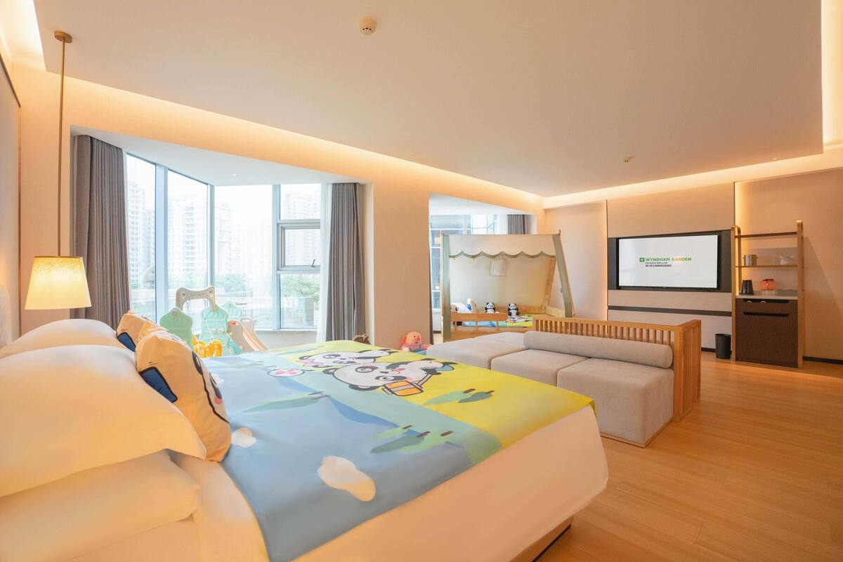 A modern hotel room with a large bed, a sofa, a TV, and a balcony with a view of buildings.