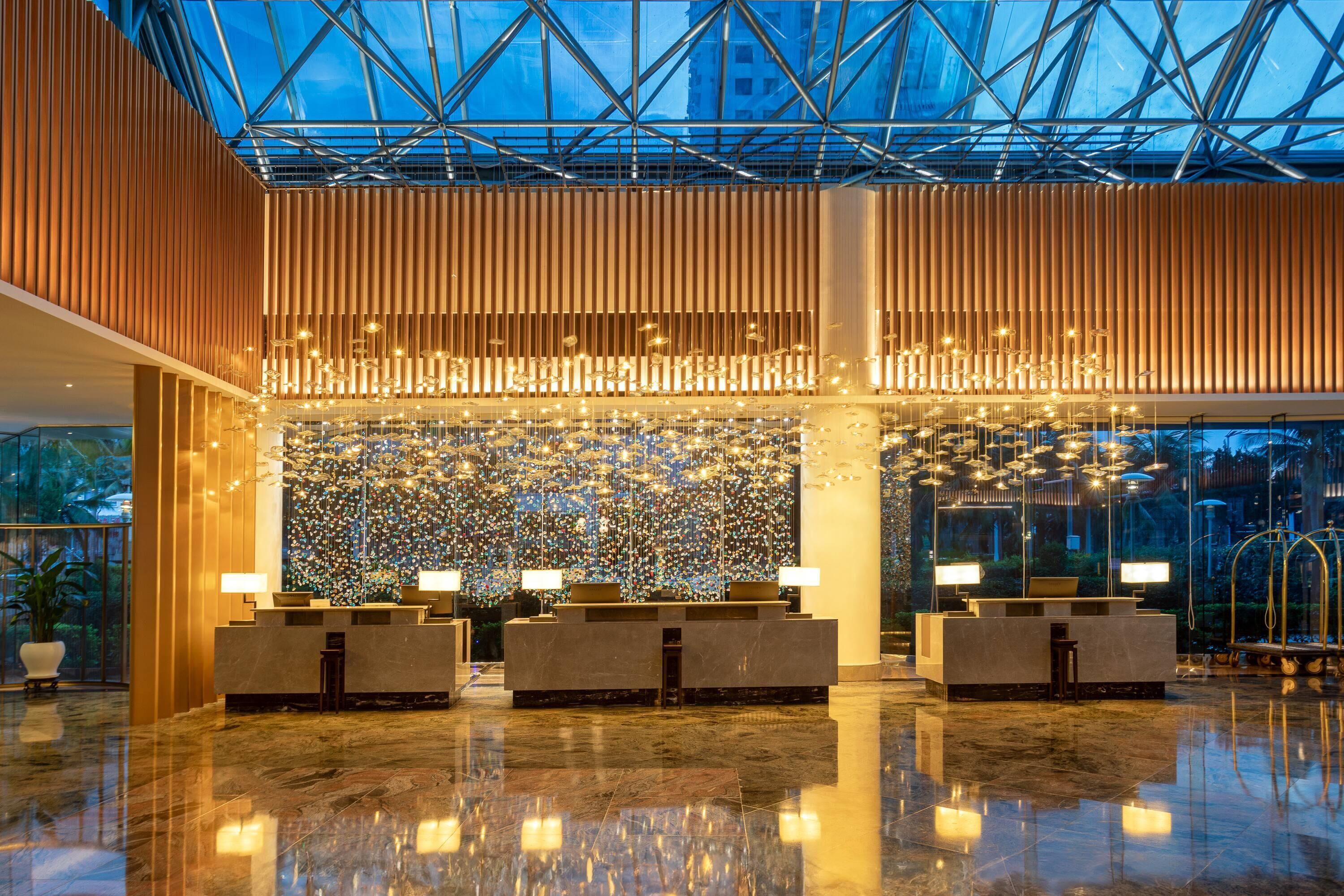 A modern hotel lobby with a glass ceiling, reflective floors, and a central decorative wall.