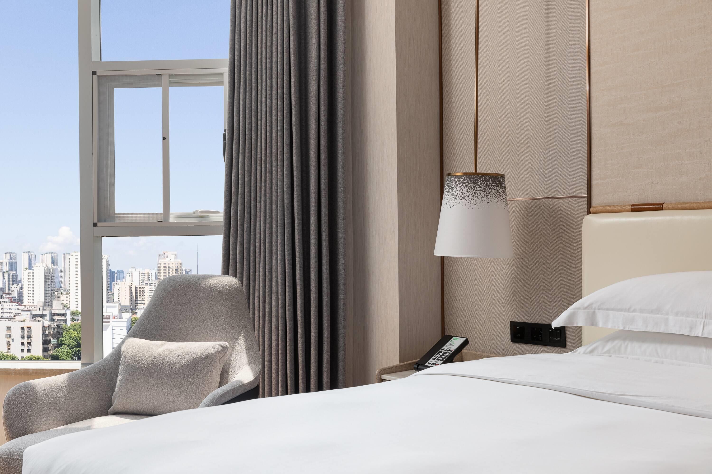 Habitación de hotel con una cama, una silla, una lámpara, un teléfono y vistas a un paisaje urbano.