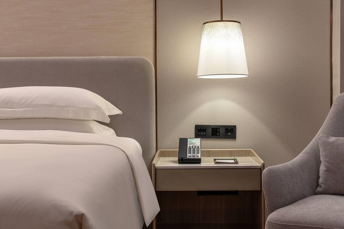 A hotel room with a bed, a bedside table with a phone, a lamp, and a chair.