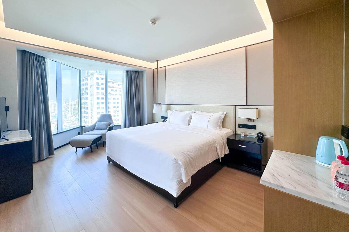 A modern hotel room with a large bed, a desk, a chair, and a view of the city.
