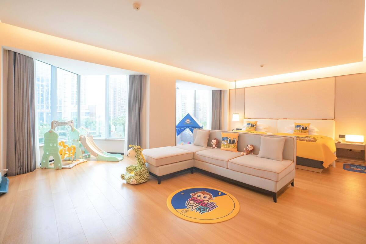A modern hotel room with a large window, a sofa, a small table, and a play area with toys.