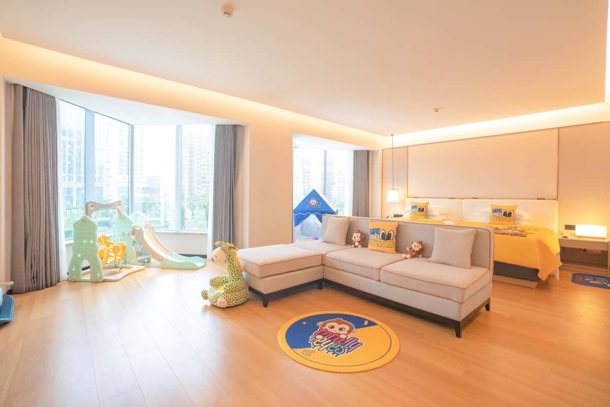A modern hotel room with a large window, a sofa, a small table, and a play area with toys.