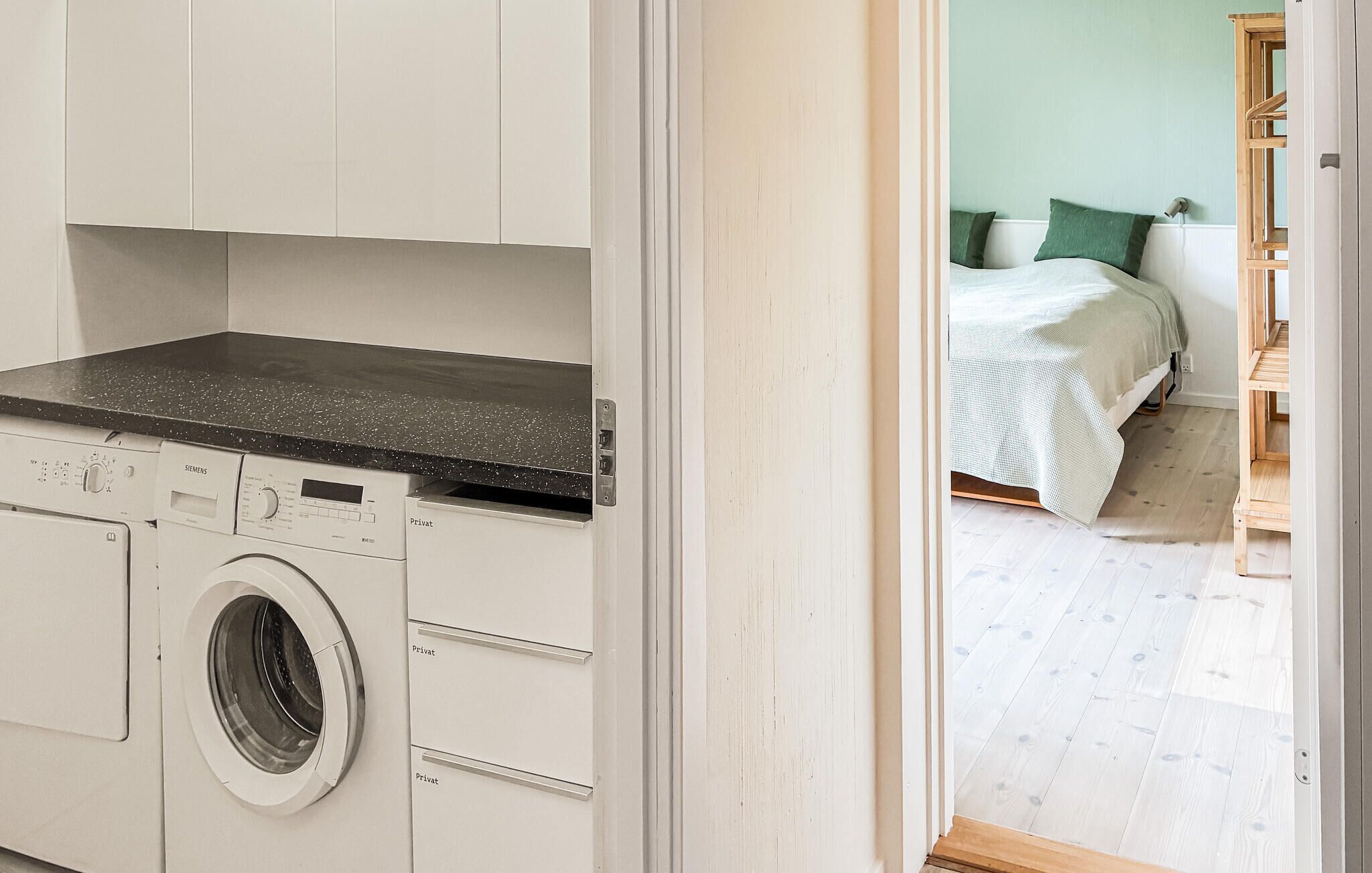 Un espace buanderie compact comprenant un lave-linge et un sèche-linge, une kitchenette avec un plan de travail noir, et un lit avec des oreillers verts dans la pièce adjacente.