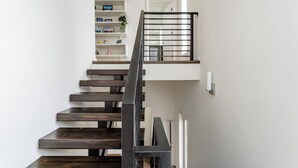 Een moderne trap met houten treden en een metalen leuning, leidend naar een overloop op de tweede verdieping met een witte muur en een plank met diverse spullen.