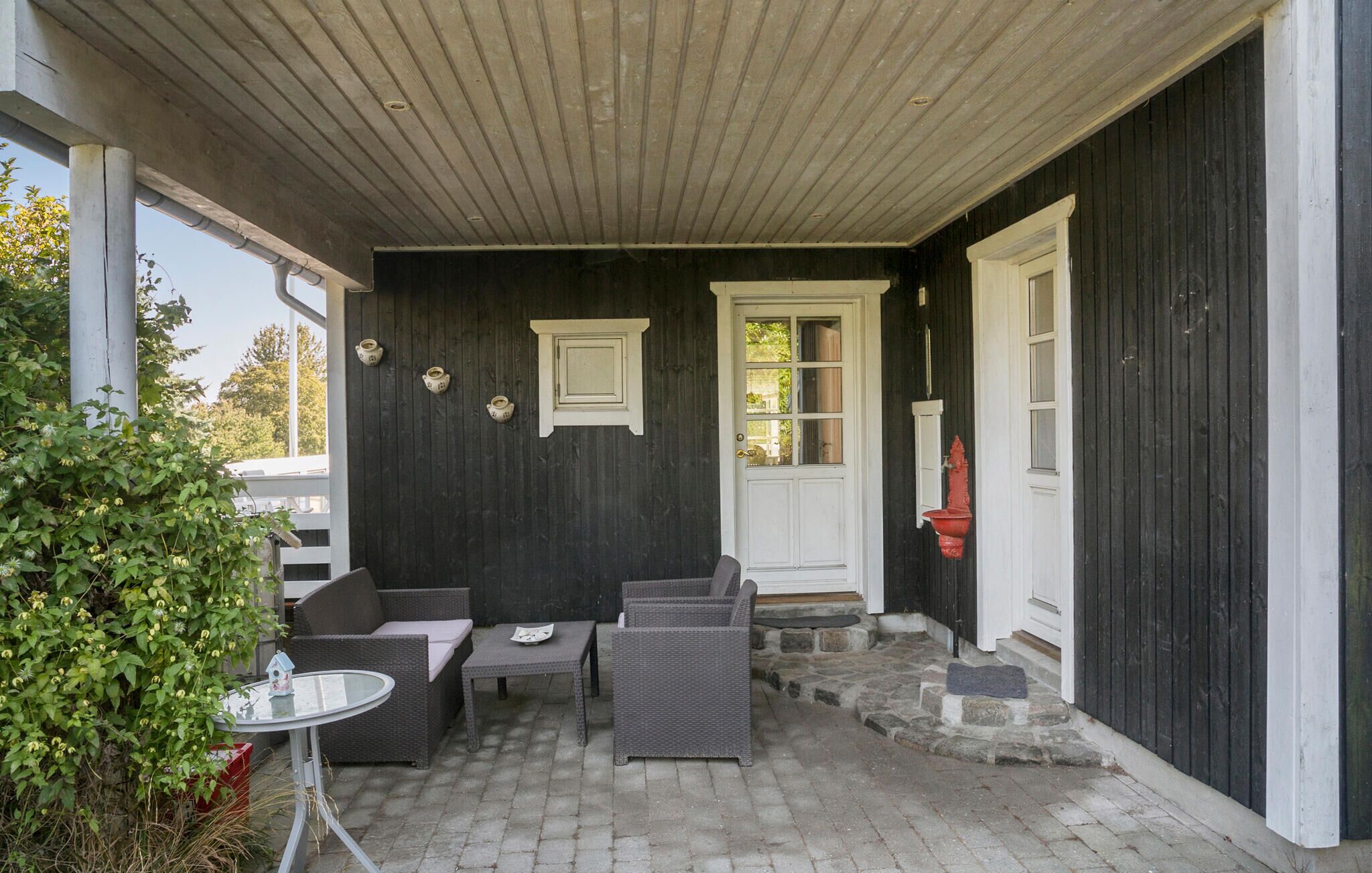 Een overdekt terras met rieten meubels en een kleine tafel, naast een donker houten gebouw met een witte deur en ramen.