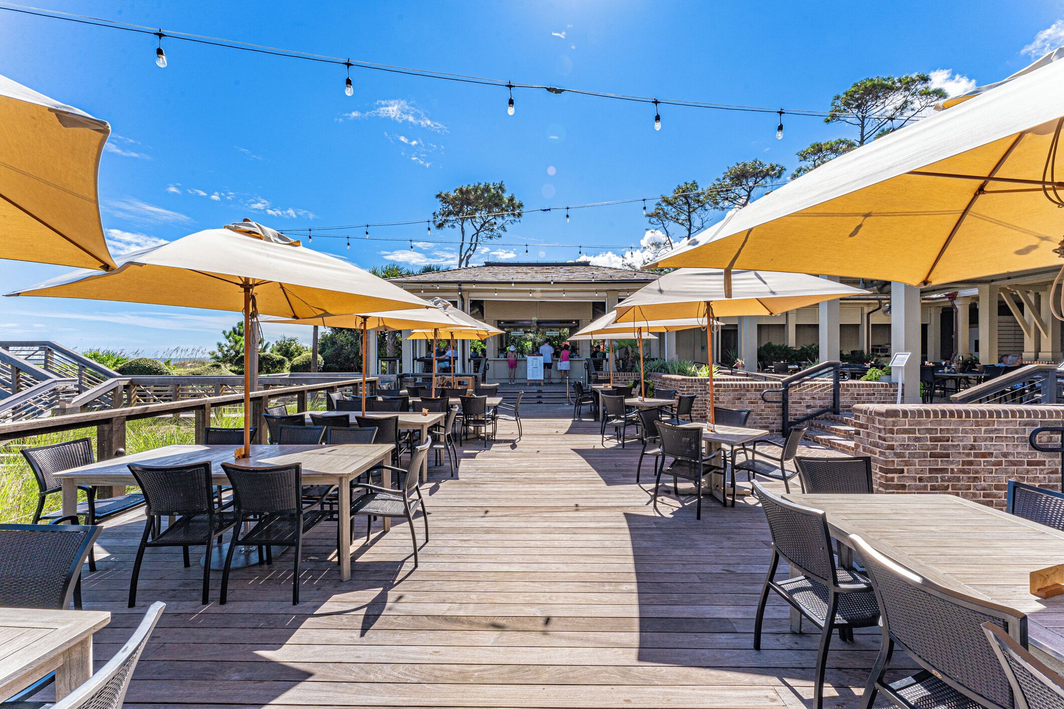 Un espace extérieur aménagé en terrasse, avec des tables et des chaises en bois, des parasols jaunes et un mur en briques.