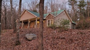 A cabin with a stone exterior and green roof, surrounded by a forest with fallen leaves.