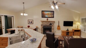 A spacious living area with a stone fireplace, a kitchen island with a sink, a dining table, and a television mounted on the wall.