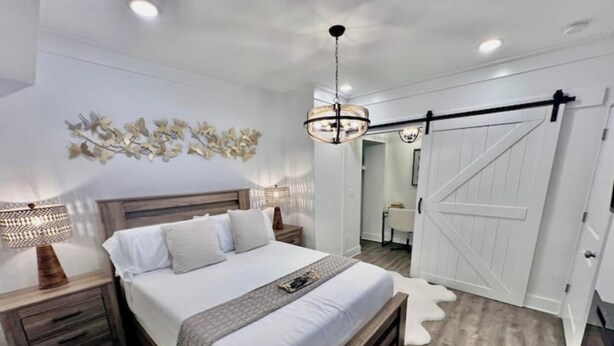 A bedroom with a bed, nightstands, a chandelier, a sliding barn door, and a wall-mounted artwork. - Glamorous Hideaway (Gainesville)