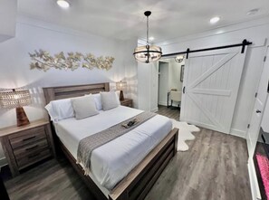 A bedroom with a bed, nightstands, a chandelier, a sliding barn door, and a wall-mounted artwork. - Glamorous Hideaway (Gainesville)