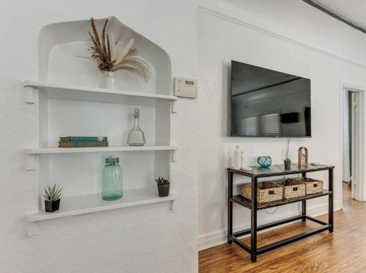 Una habitación con paredes blancas, suelo de madera, un estante blanco con adornos, una consola negra con cestas y un televisor de pantalla plana montado en la pared.