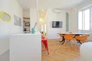 A modern kitchen with a microwave, a dining area with a table and chairs, a TV mounted on the wall, and a large window.