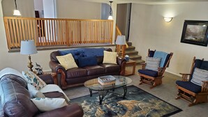 Uma sala de estar com um sofá de couro marrom, uma mesa de centro de vidro, cadeiras de madeira e uma escada ao fundo.