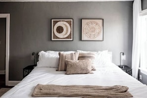 Un lit bien fait, avec du linge de lit blanc et plusieurs oreillers, une couverture en tissu et deux œuvres d’art encadrées accrochées au mur au-dessus du lit.