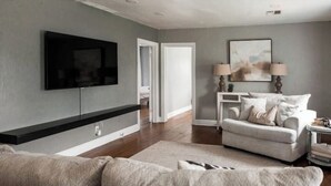 A modern living room with a flat-screen TV mounted on the wall, a grey sofa, a white armchair, and a small side table with lamps. - 7 min to Downtown / W/D / Historic / Central (Oklahoma City)