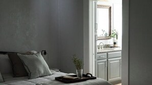 Une chambre à coucher avec un lit, des oreillers, une table de chevet avec un plateau, et une vue sur une cuisine et une salle de bain par une porte ouverte.