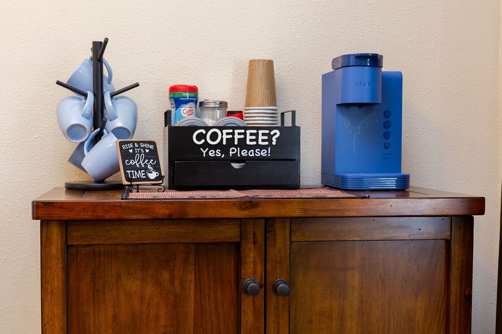 A coffee station with a coffee maker, a coffee dispenser, and a sign that says "COFFEE? Yes, please!".