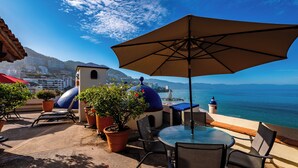 Eine Terrasse mit Tisch und Stühlen, einem großen Sonnenschirm, Kübelpflanzen und Blick auf das Meer und Gebäude im Hintergrund.