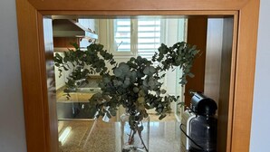 Une cuisine avec un miroir à cadre en bois au-dessus d’un plan de travail en granit, un vase en verre contenant des feuilles vertes et une machine à café.