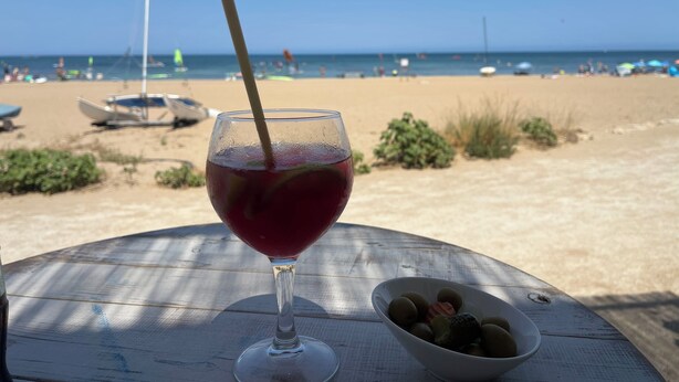 Un verre d’une boisson rouge est posé sur une table en bois, à côté d’un bol d’olives, le tout sur fond de plage.