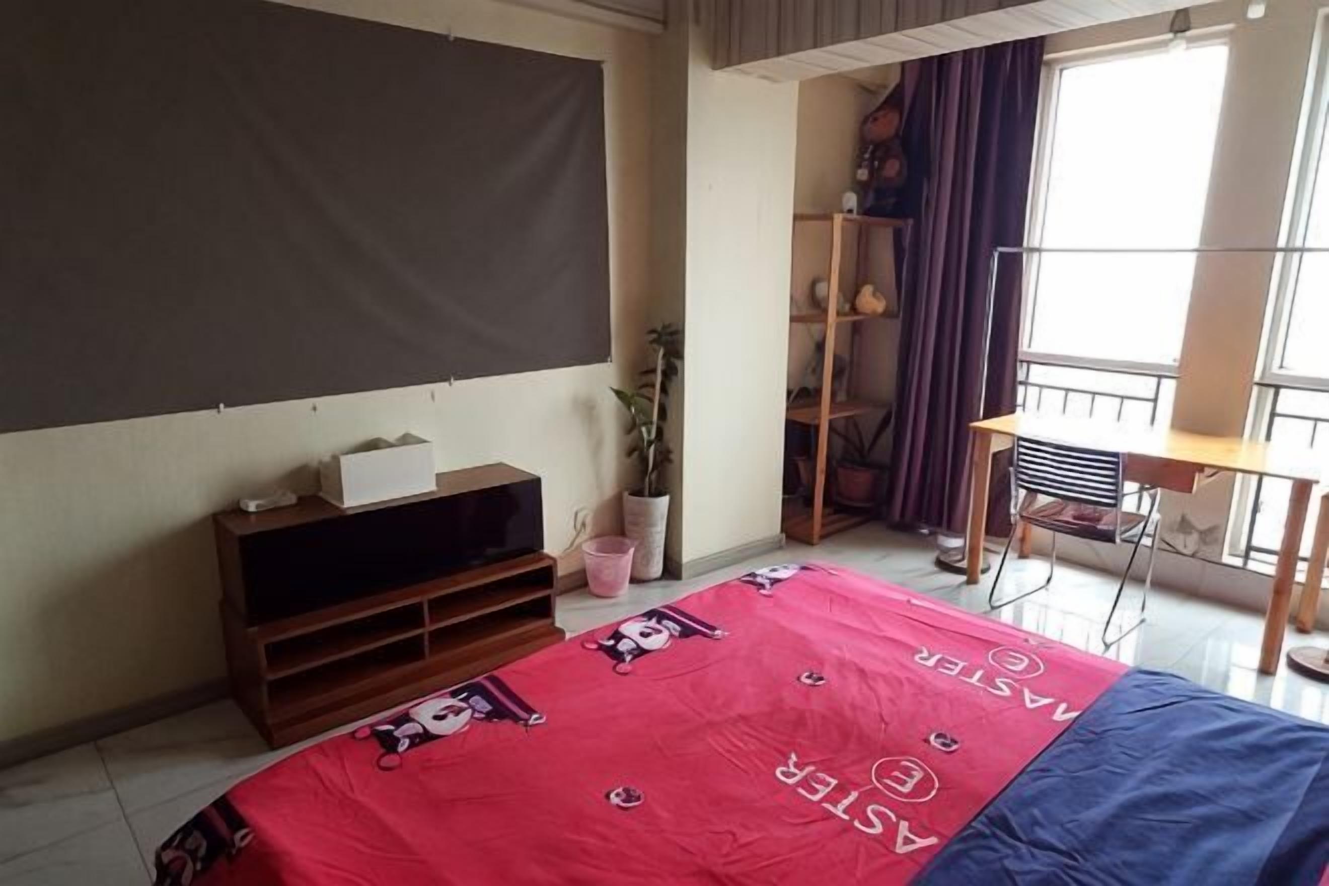 Une chambre à coucher comprenant un lit, un téléviseur posé sur un meuble, un bureau, une chaise, une fenêtre avec des rideaux et une étagère ornée d’objets décoratifs.