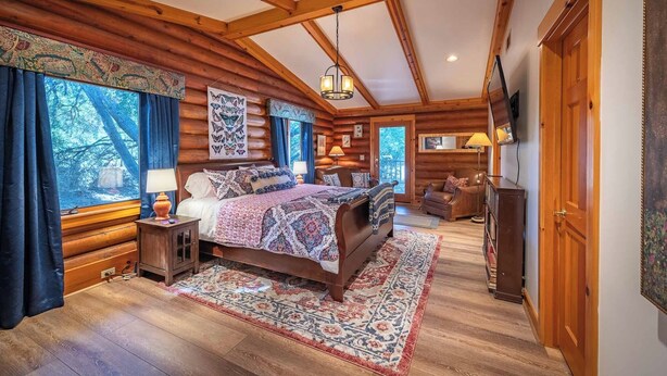 A log cabin bedroom with a large bed, wooden beams, and a view of trees through the window.