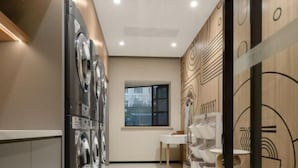 A modern laundry room with stacked washing machines, a sink area, and a window providing a view of the cityscape. - Home Inn (Shanghai Stadium Wanping South Road) (Shanghai)