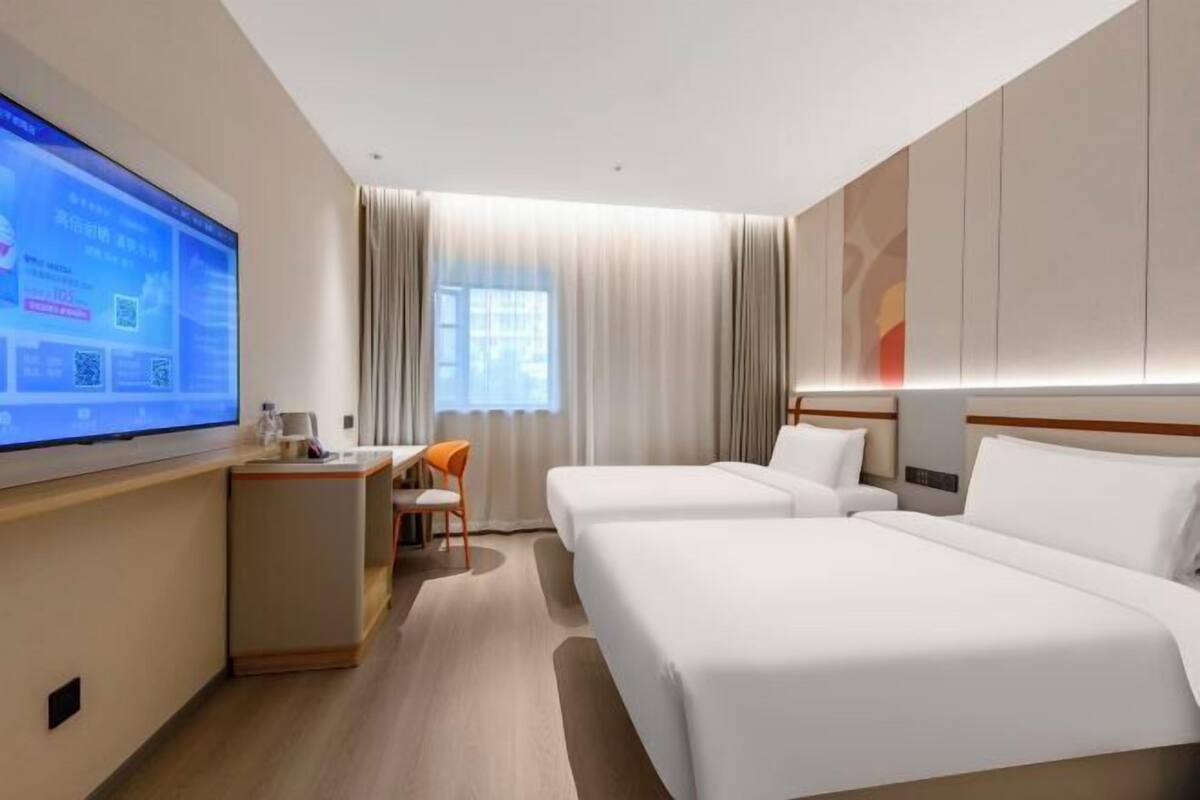 Ein Hotelzimmer mit zwei Betten, einem Schreibtisch mit Stuhl, einem Wandfernseher und einem Fenster mit Vorhängen.