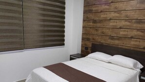 A bedroom with a bed, a nightstand, a window with blinds, and a wooden wall panel.