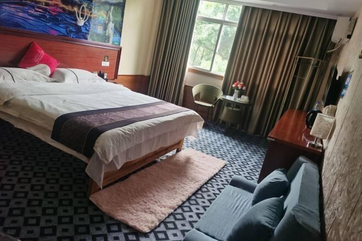 A hotel room with a large bed, a small table with a chair, and a view of the outdoors through a window.
