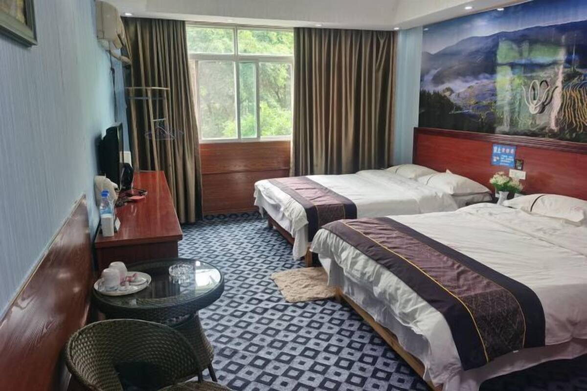 A hotel room with two beds, a small table, a chair, a window with curtains, and a painting on the wall.